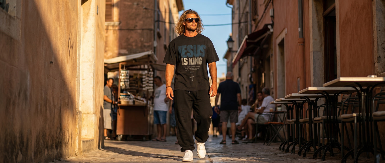 Man in a faded JESUS IS KING tee walking a narrow old-town street at sunset