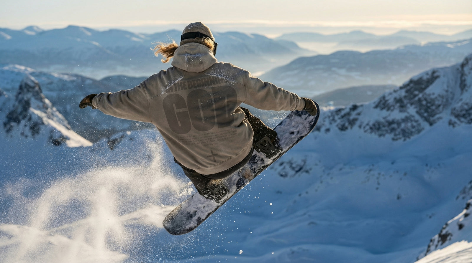 Snowboarder mid-air wearing a sand IN THE BEGINNING GOD hoodie above snowy peaks